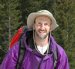 Photo of David Cook coming out of the woods, wearing a hat, jacket and backpack.